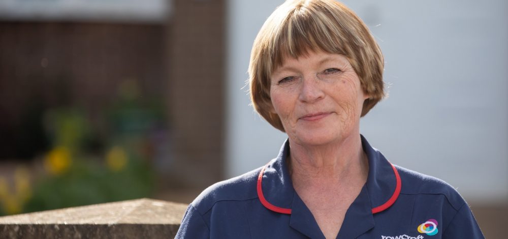 A clinical nurse in a blue uniform smiles ouotdoors.