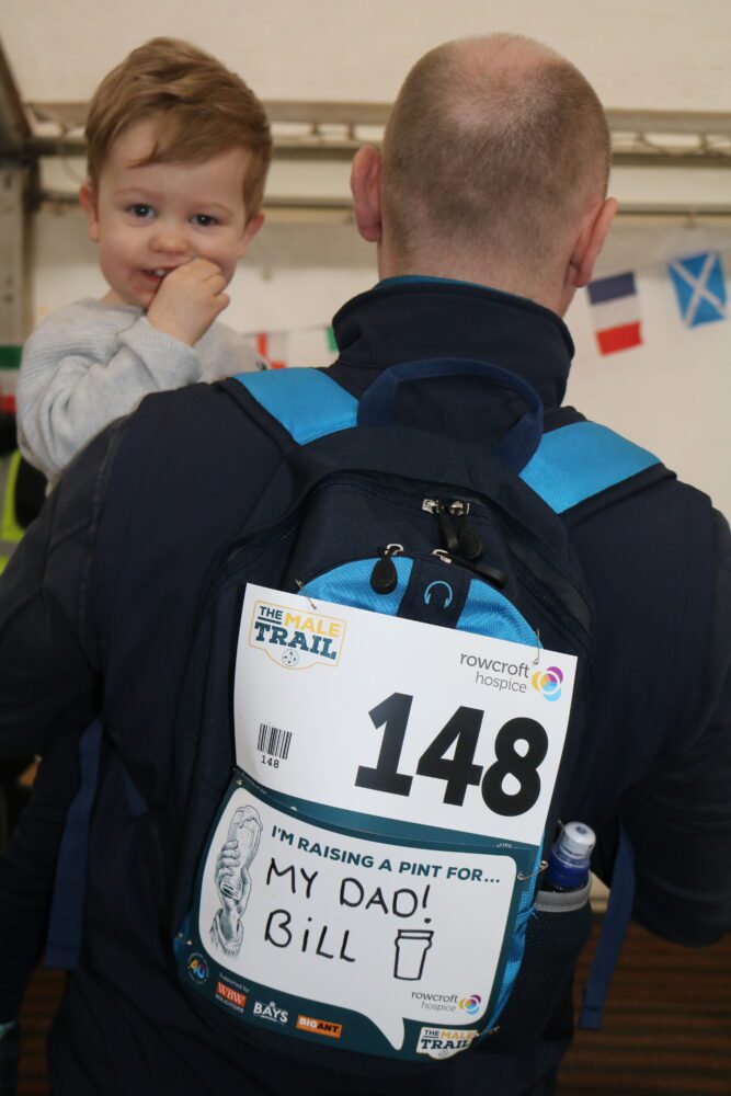 Rob Burkitt holding a baby facing backwards wearing his Male Trail participant number and dedication. The dedication reads 'My dad Bill'. 