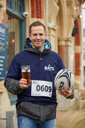 A man stands, holding a pint of Bays and a rugby ball