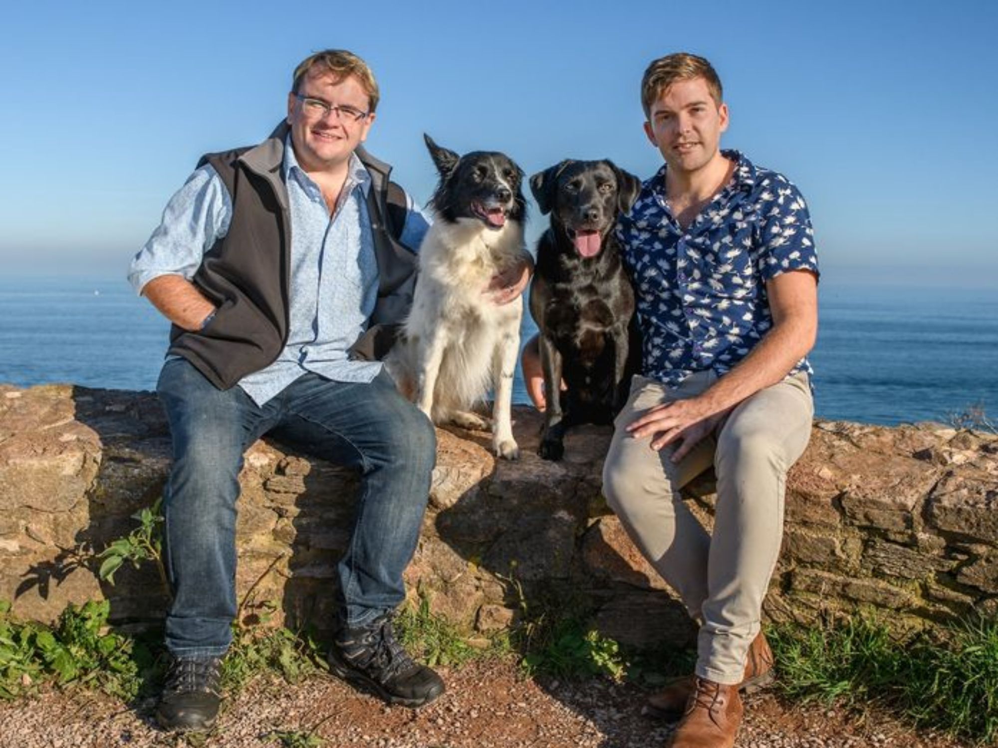 Rowcroft Nurse, Kieron, and husband sat on a wall with their two dogs and the sea behind them.